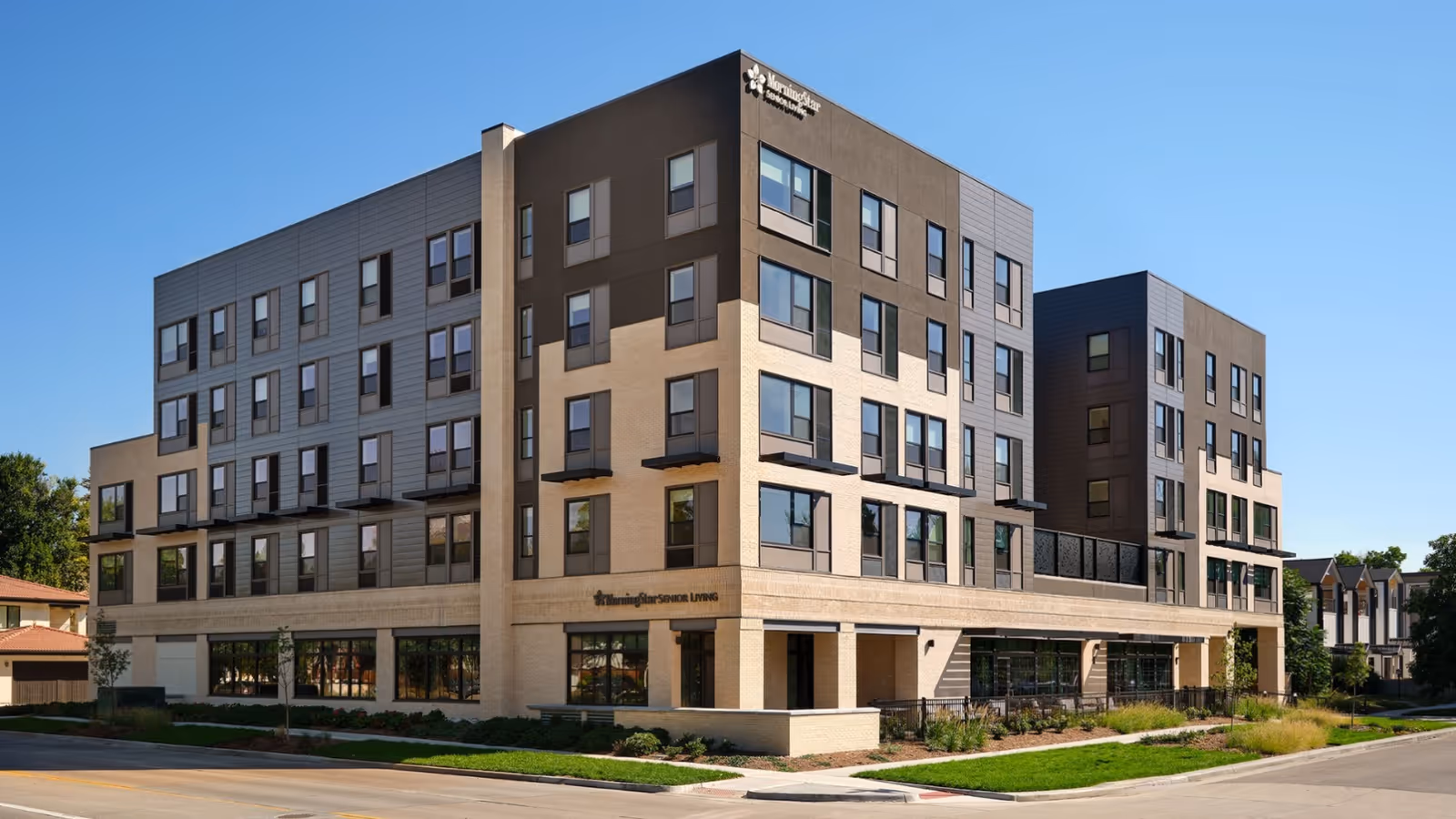 Exterior front view of a modern multi-story assisted living and memory care building with numerous windows and landscaped grounds under a clear blue sky.