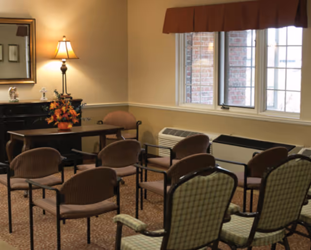 A small meeting or activity room with several chairs arranged in rows facing a wooden table with a flower arrangement. The room has beige walls, a window with a brown valance, a lamp on a sideboard, and a mirror hanging on the wall.
