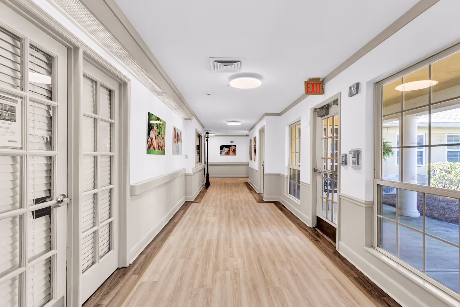 A bright, clean hallway in an assisted living facility with light wood flooring, white walls with beige wainscoting, and several framed pictures on the walls. There are large windows on the right side letting in natural light and a glass door with an exit sign above it. On the left side, there are double doors with glass panes and white blinds.