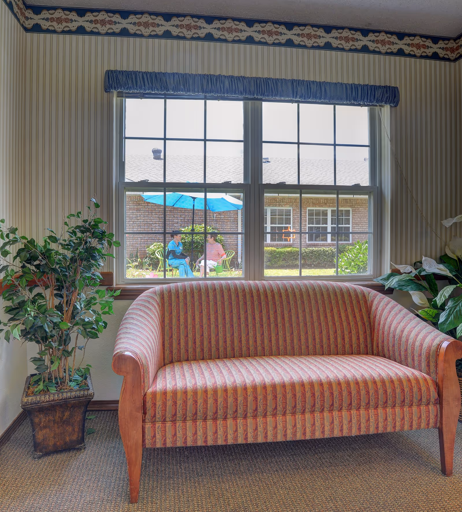 A striped upholstered loveseat sits before a large window that looks out onto a courtyard where two people sit under a blue umbrella.
