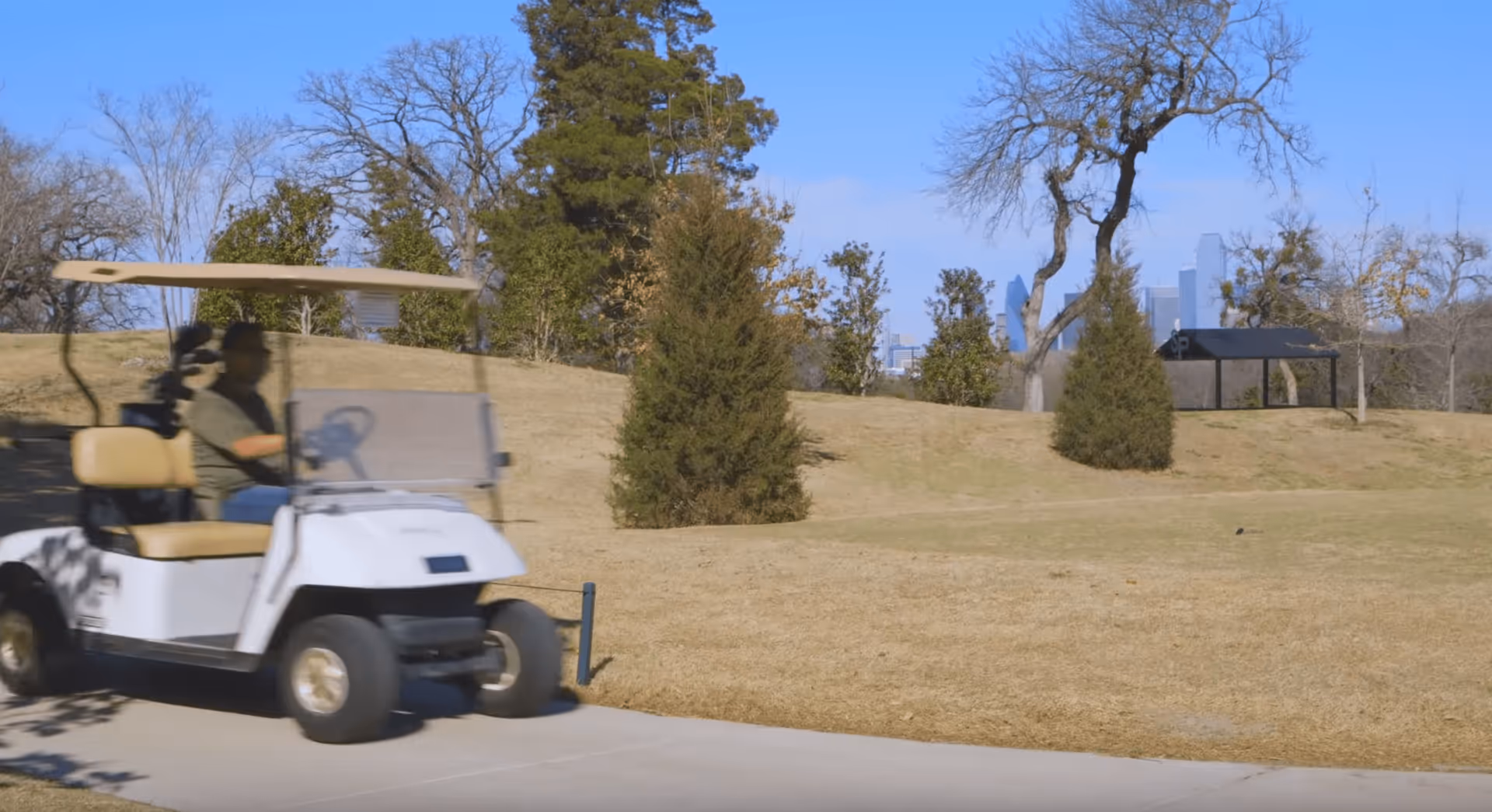 A person driving a white golf cart on a paved path in a park-like outdoor area with grass, trees, and a small pavilion in the background under a clear blue sky.