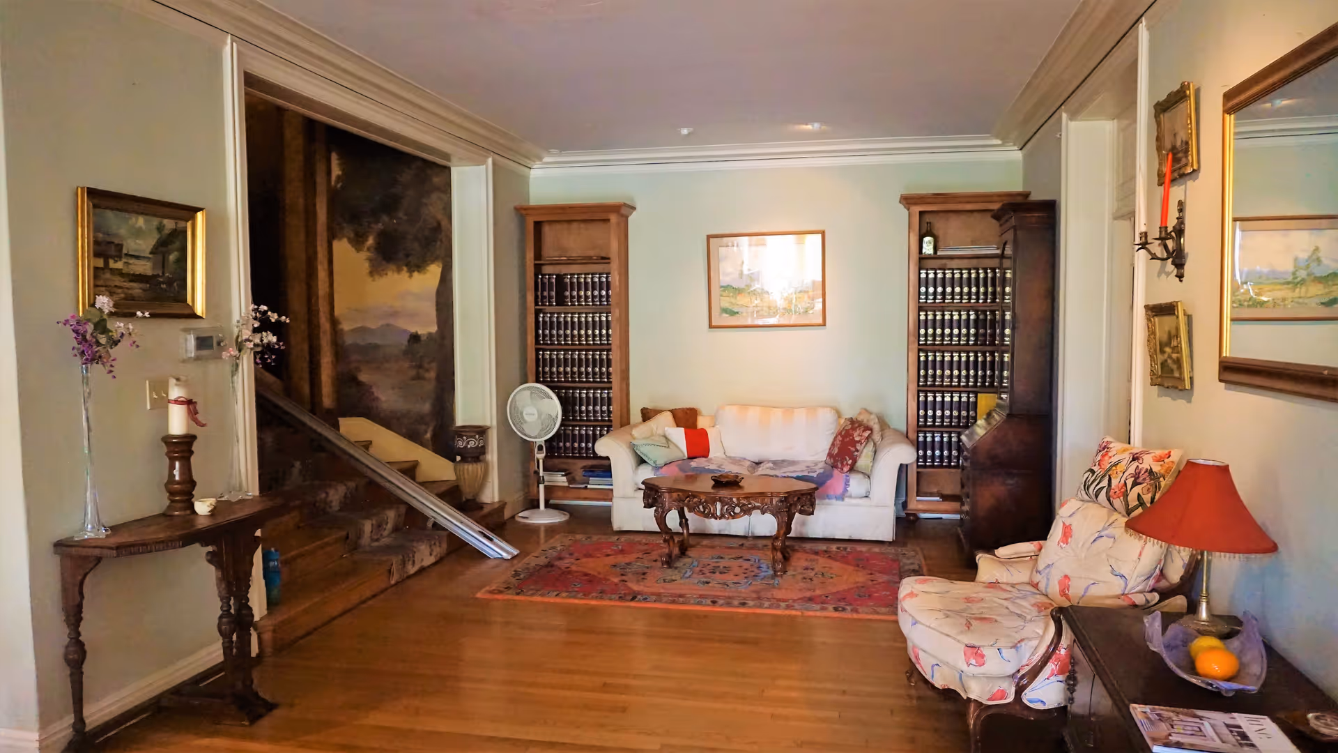 Cozy living room with sofas and armchairs around a coffee table on a rug, flanked by bookshelves and a staircase.