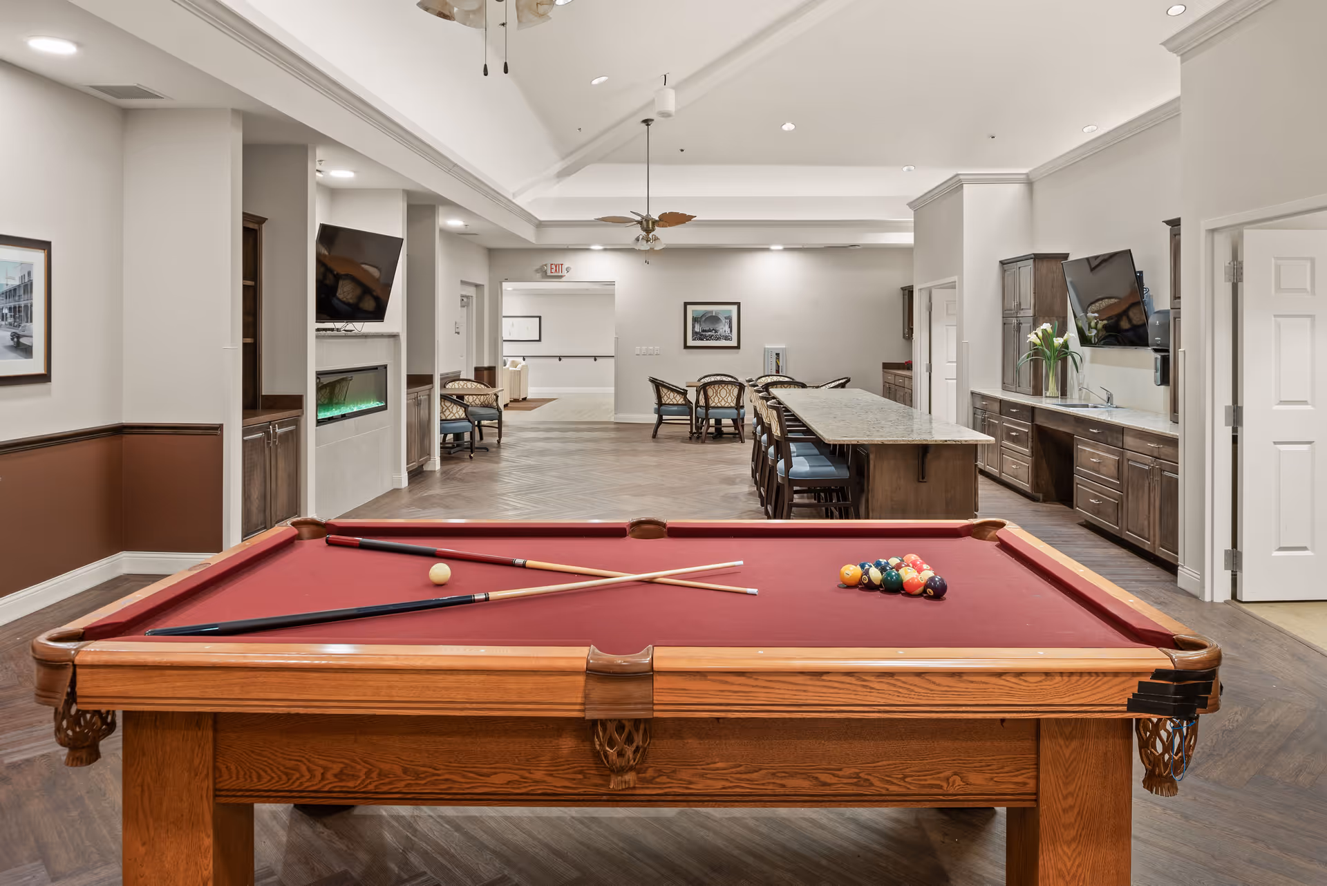 Interior view of a senior living facility common area featuring a red felt pool table with cues and balls arranged for a game. In the background, there is a long granite countertop with bar stools, several chairs and tables, a wall-mounted TV, and a modern electric fireplace. The room has wood flooring, neutral-colored walls, and recessed lighting.