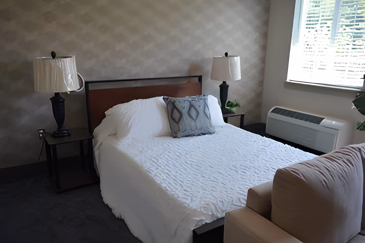 A bedroom with a neatly made bed covered in a white textured bedspread and a decorative gray pillow. There are two black bedside tables, each with a black lamp with white lampshades. A window with blinds is on the right side, allowing natural light into the room. A beige armchair is partially visible in the foreground.