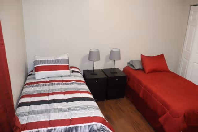 A small bedroom with two single beds separated by two black nightstands, each with a gray lamp. One bed has a striped comforter in red, white, gray, and black, while the other bed has a solid red comforter with a gray pillow. The room has wooden flooring and a white wall with a closed white door on the right side.