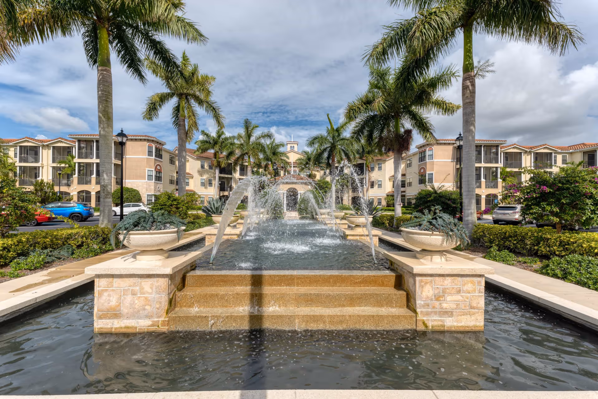 Outdoor view of Addington Place of Jupiter featuring a large rectangular water fountain with multiple water jets, surrounded by palm trees and landscaped greenery. The multi-story residential building with balconies and a tiled roof is visible in the background under a partly cloudy sky.