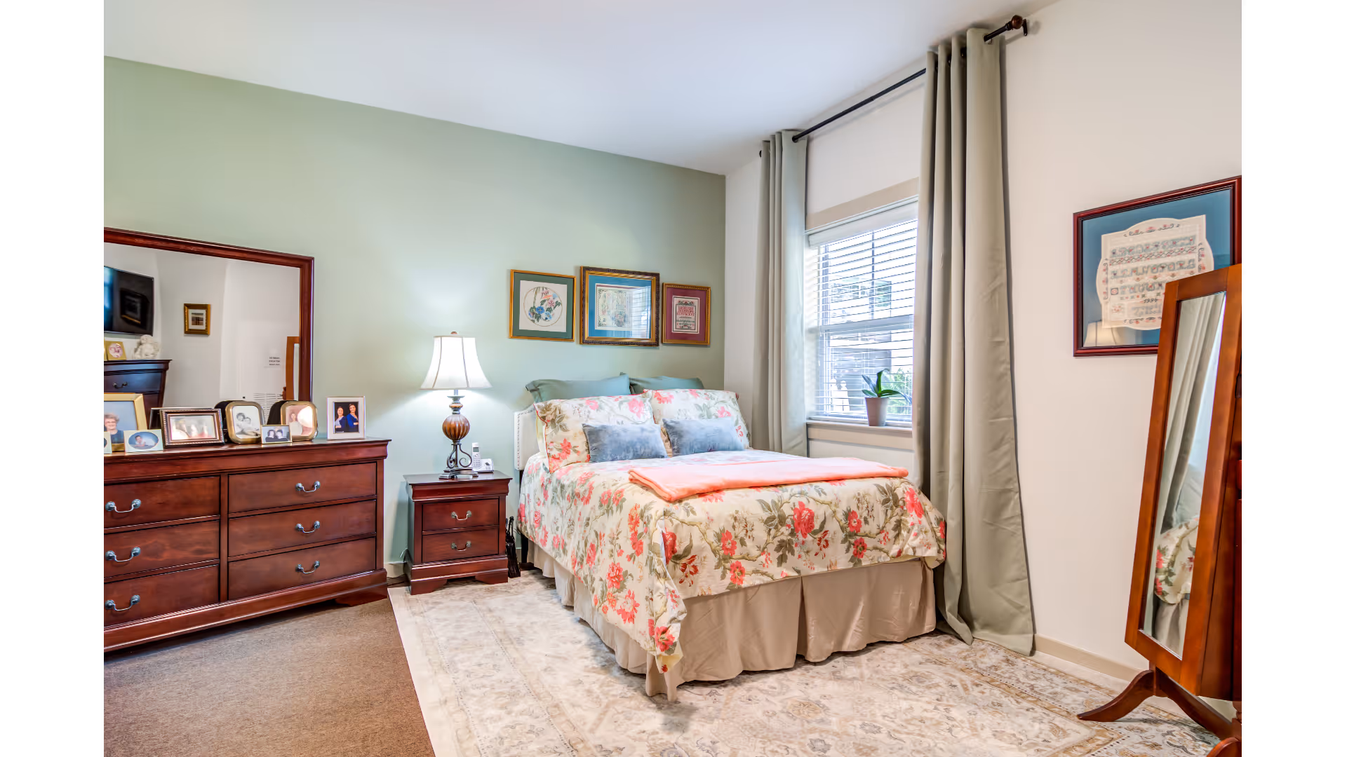 A cozy bedroom with a floral-patterned bedspread on a double bed, a wooden dresser with a large mirror and framed photos, a nightstand with a lamp, a window with beige curtains, a framed picture on the wall, and a standing mirror.