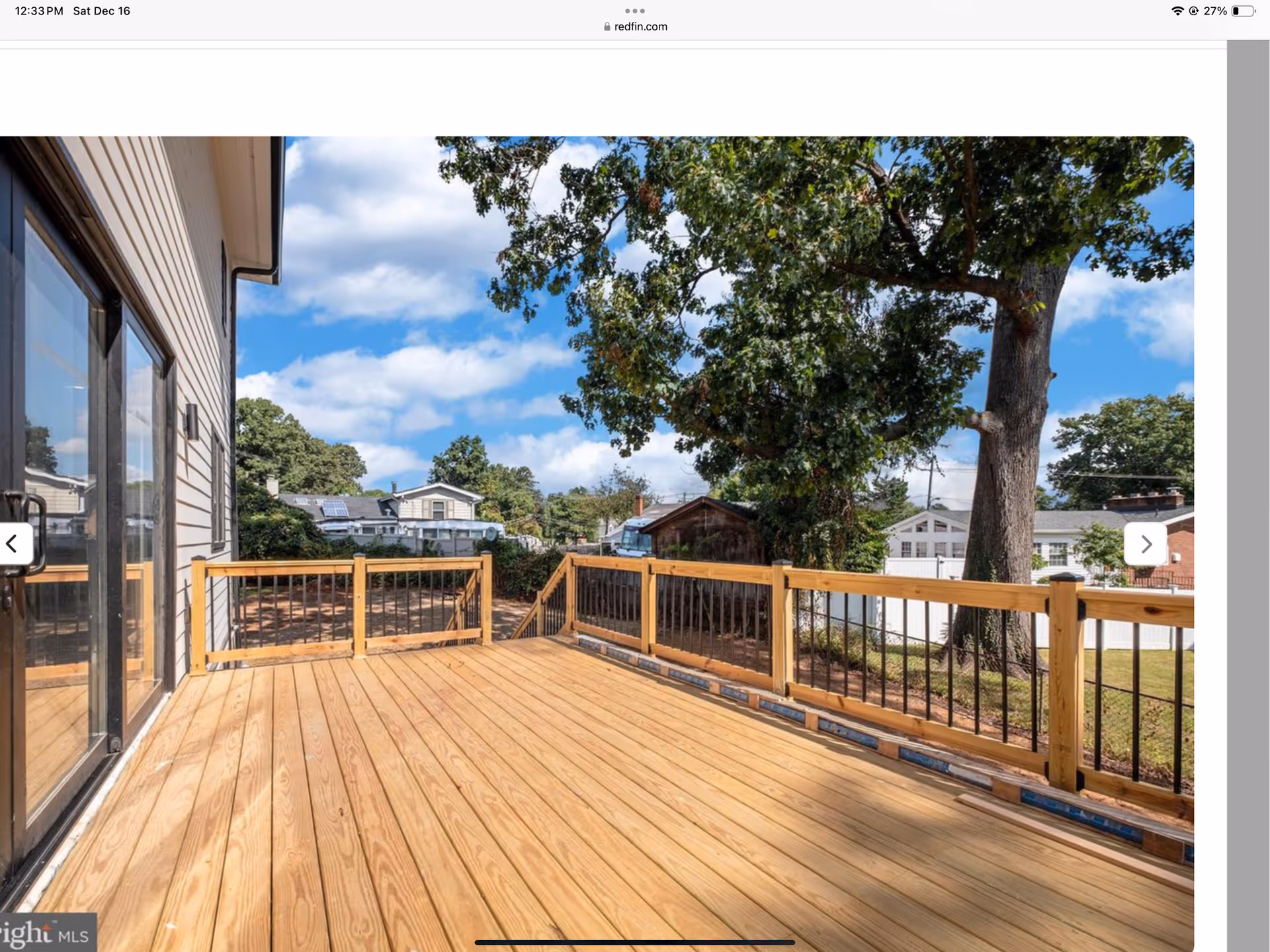 A spacious wooden deck attached to a house with sliding glass doors, overlooking a backyard with trees and neighboring houses under a partly cloudy sky.