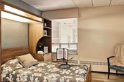 A small, neatly arranged bedroom in a senior living facility featuring a single bed with floral bedding, a wooden headboard with built-in shelves, a window with blinds, a small table with a menu or brochure, and a chair with a cushion.