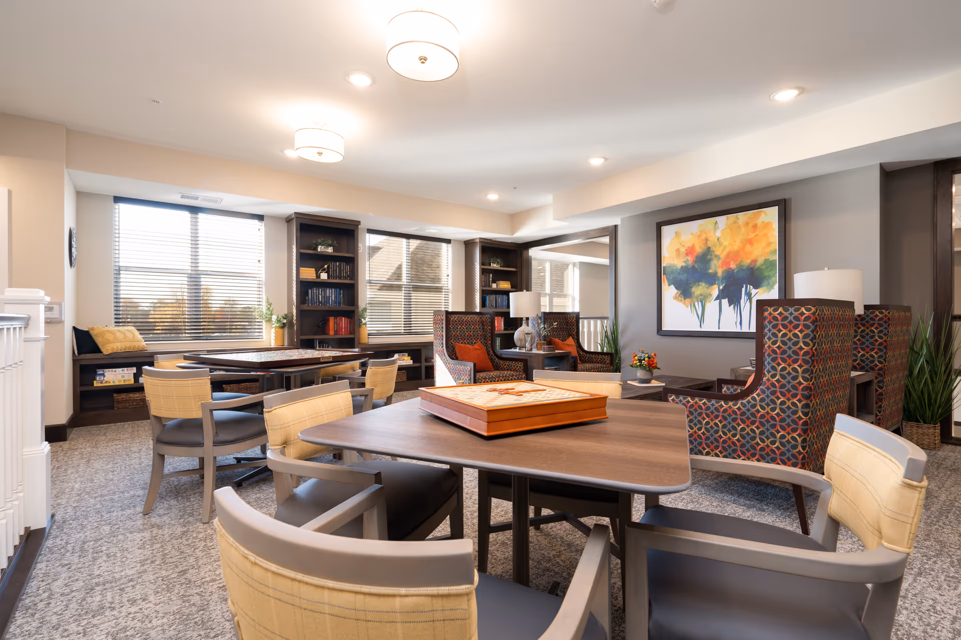 A bright and spacious common area in a senior living facility featuring multiple tables with chairs arranged around them. There are two patterned armchairs with orange cushions near a side table with a lamp and a colorful abstract painting on the wall. Large windows with blinds allow natural light to fill the room, and built-in shelves hold books and decorative plants. The carpeted floor and soft lighting create a warm and inviting atmosphere.