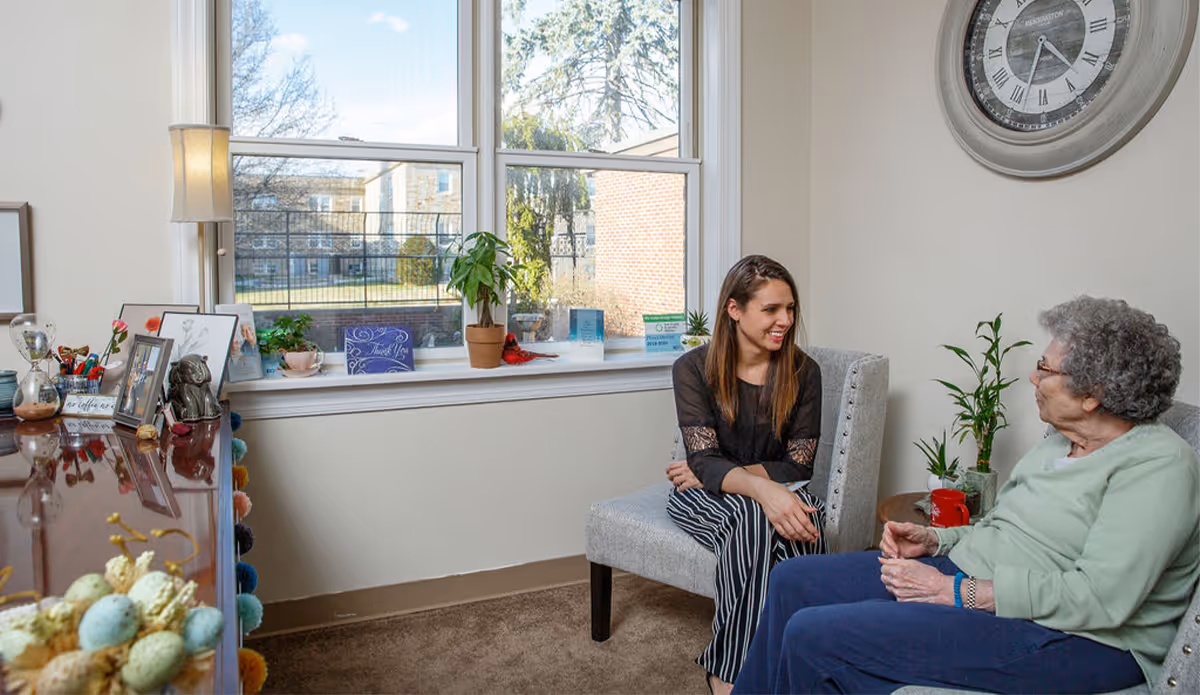 A young woman and an elderly woman sitting and talking in a cozy room with a large window, a clock on the wall, and various plants and decorations on a dresser and windowsill.