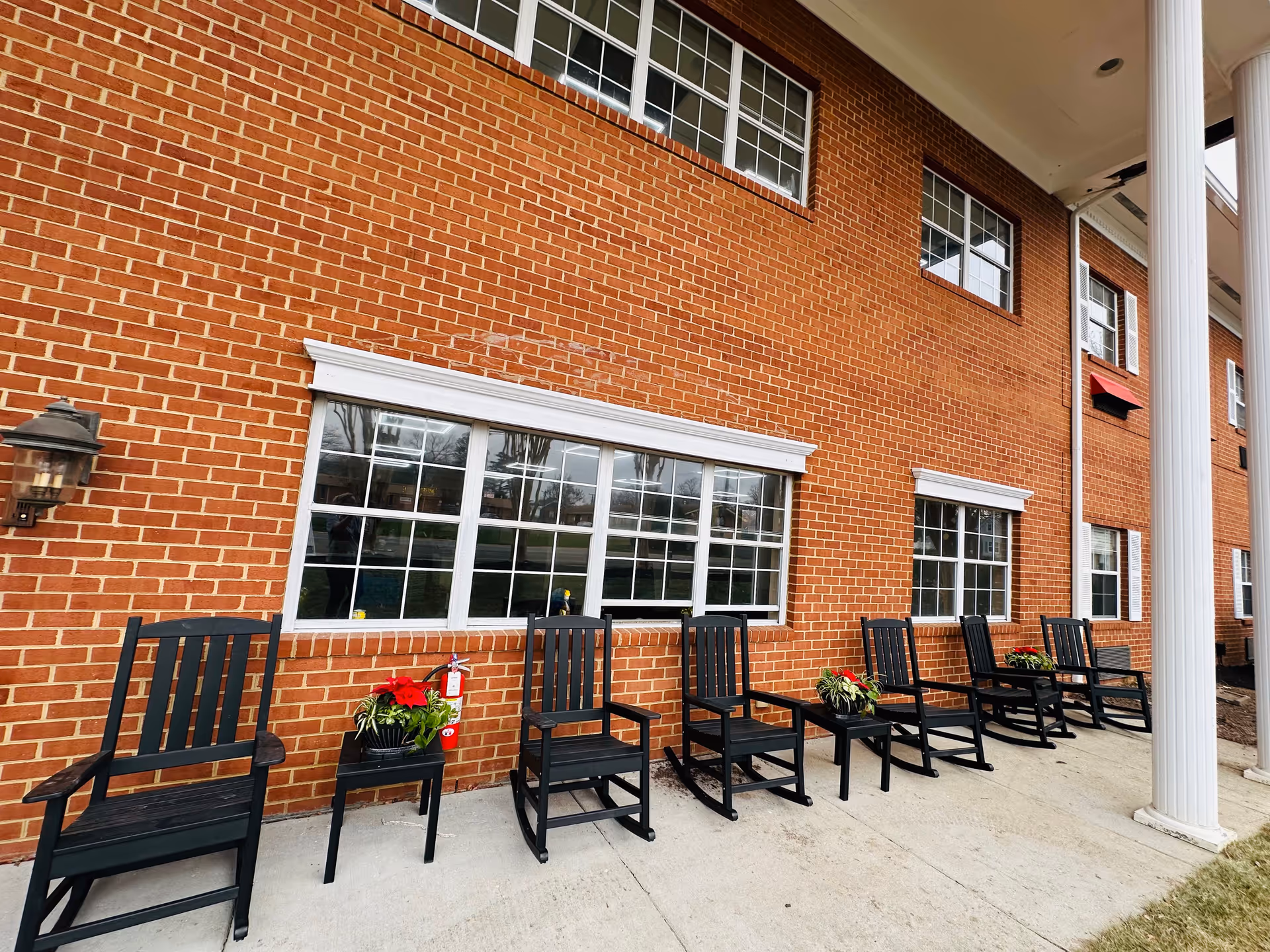 Brick building front with a row of black rocking chairs and potted plants beneath large windows and white columns.