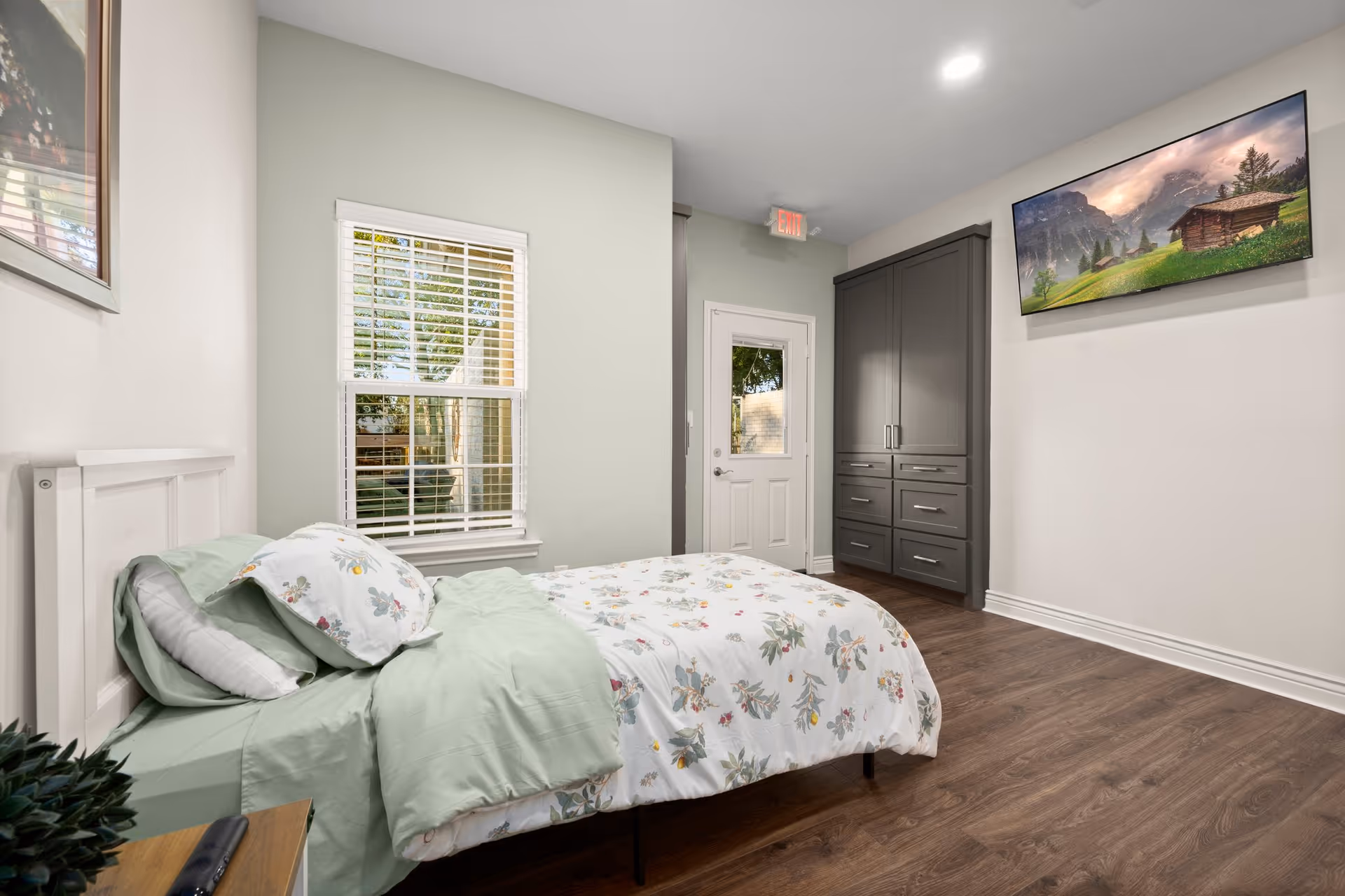 Bright assisted-living bedroom with a single bed dressed in floral bedding, a window with blinds, a gray wardrobe, wall-mounted TV, and a door.
