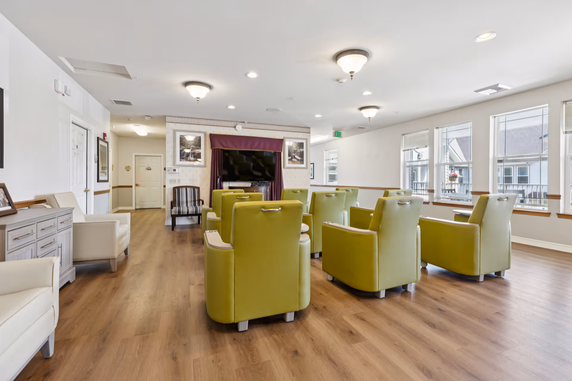A bright and spacious common living area with wooden flooring, featuring a group of green armchairs arranged in rows facing a large flat-screen TV mounted on a wall with framed pictures on either side. The room has large windows allowing natural light to fill the space, white walls, and ceiling lights. Additional seating includes beige armchairs and a wooden sideboard along one wall.