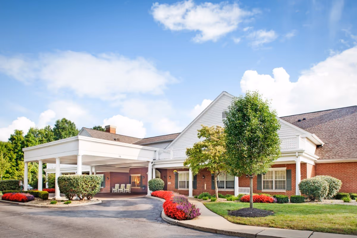 Front exterior of a red-brick senior living building with a covered entrance, driveway and landscaped flower beds.
