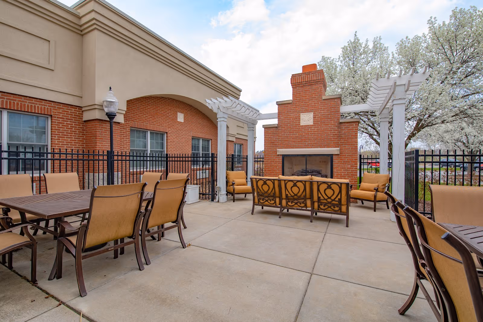 Outdoor patio area with cushioned chairs and tables arranged around a brick fireplace. The patio is enclosed by a black metal fence and has white pergola structures. Trees with white blossoms are visible in the background under a partly cloudy sky.