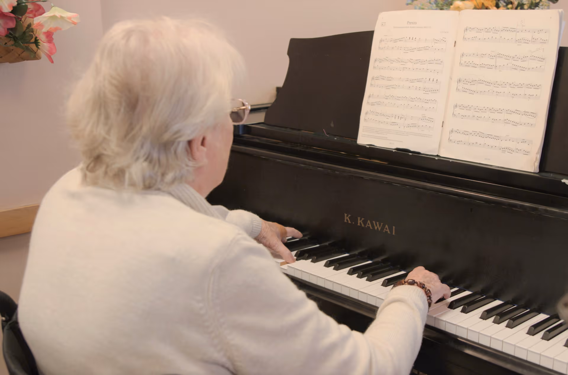 An elderly person with white hair and glasses playing a black K. Kawai piano with sheet music placed on the music stand. There is a flower arrangement visible on the left side.