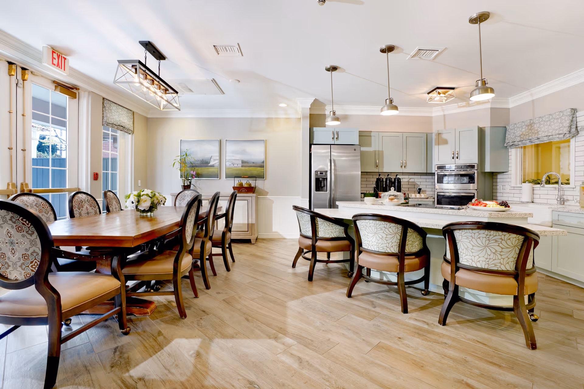 A bright and spacious kitchen and dining area with a large wooden dining table surrounded by upholstered chairs on the left and a kitchen island with four cushioned chairs on the right. The kitchen features light green cabinets, stainless steel appliances, pendant lighting, and a window with a patterned valance. The floor is light wood, and there are two framed landscape paintings on the far wall.