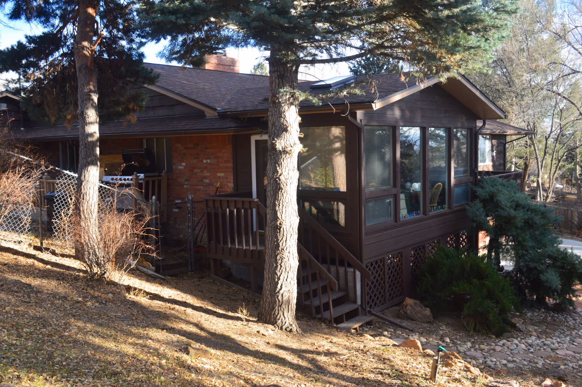 Exterior view of a single-story assisted living facility building with brown wooden siding and a brick section. The building has large windows, a small wooden staircase leading to a door, and is surrounded by trees and shrubs on a sloped, leaf-covered ground.