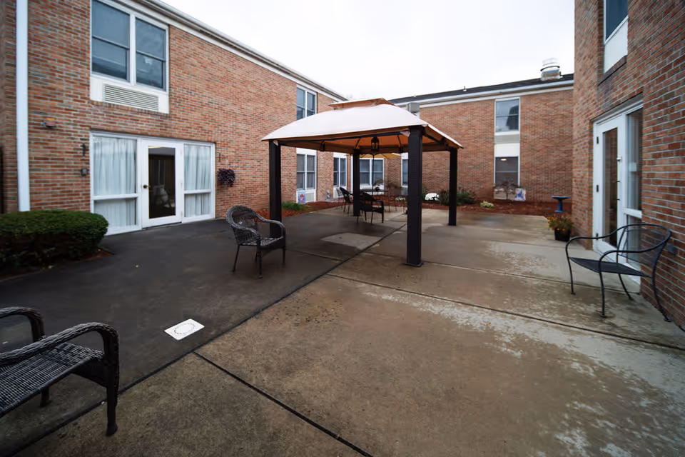 Outdoor courtyard area of a senior living facility with brick walls surrounding the space. There is a gazebo with a canopy in the center, several black metal and wicker chairs arranged around the courtyard, and some small plants along the building walls.