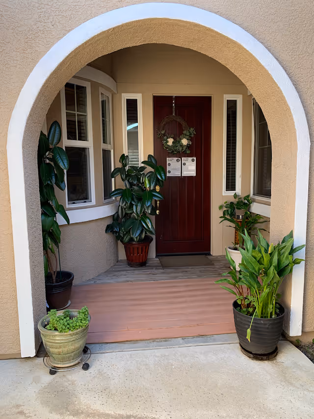 Entrance to a senior care facility with a wooden door decorated with a floral wreath and notices. The entrance is framed by a beige stucco archway and flanked by several potted green plants on either side. There are windows on both sides of the door.