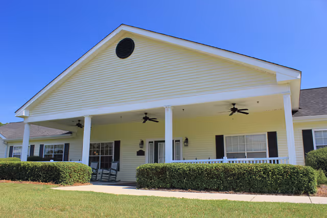 Front exterior of a yellow single-story building with a covered porch, white columns, ceiling fans, rocking chairs and trimmed hedges.