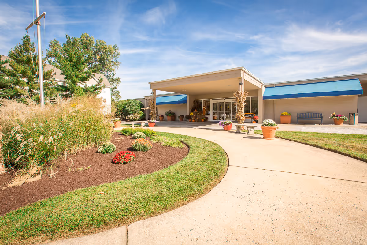 Entrance to Mallard Bay Nursing and Rehab facility with a covered drop-off area, potted plants, benches, and a landscaped garden with grass, shrubs, and flowers under a clear blue sky.