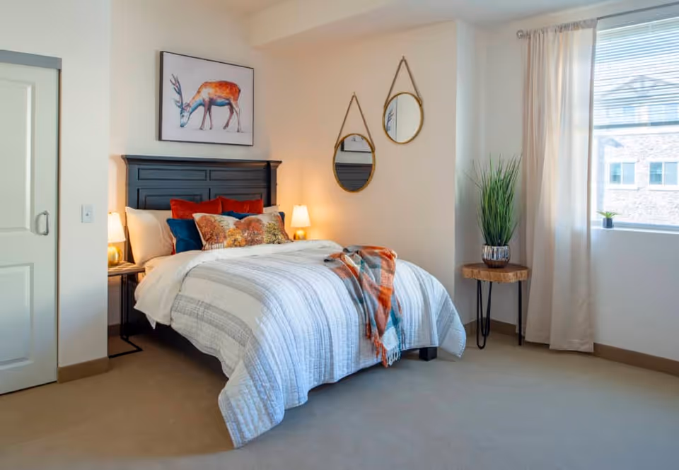 A cozy bedroom with a neatly made bed featuring a black wooden headboard, white and gray striped bedding, and multiple decorative pillows in red, blue, and floral patterns. Above the bed is a framed picture of a deer. Two round mirrors with leather straps hang on the adjacent wall. A small wooden side table with a potted plant sits near a window with white curtains, allowing natural light to fill the room.