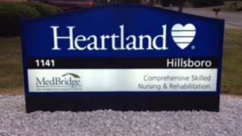 A blue Heartland Hillsboro sign with MedBridge logo stands outdoors on gravel.