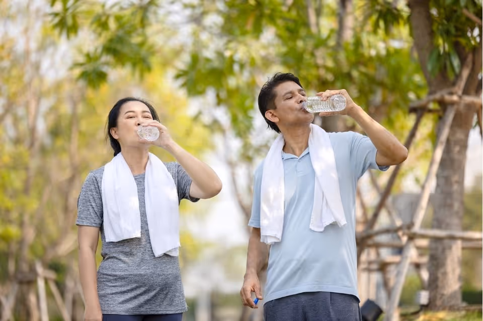 A man and a woman standing outdoors with white towels around their necks, drinking water from plastic bottles, surrounded by trees and greenery.