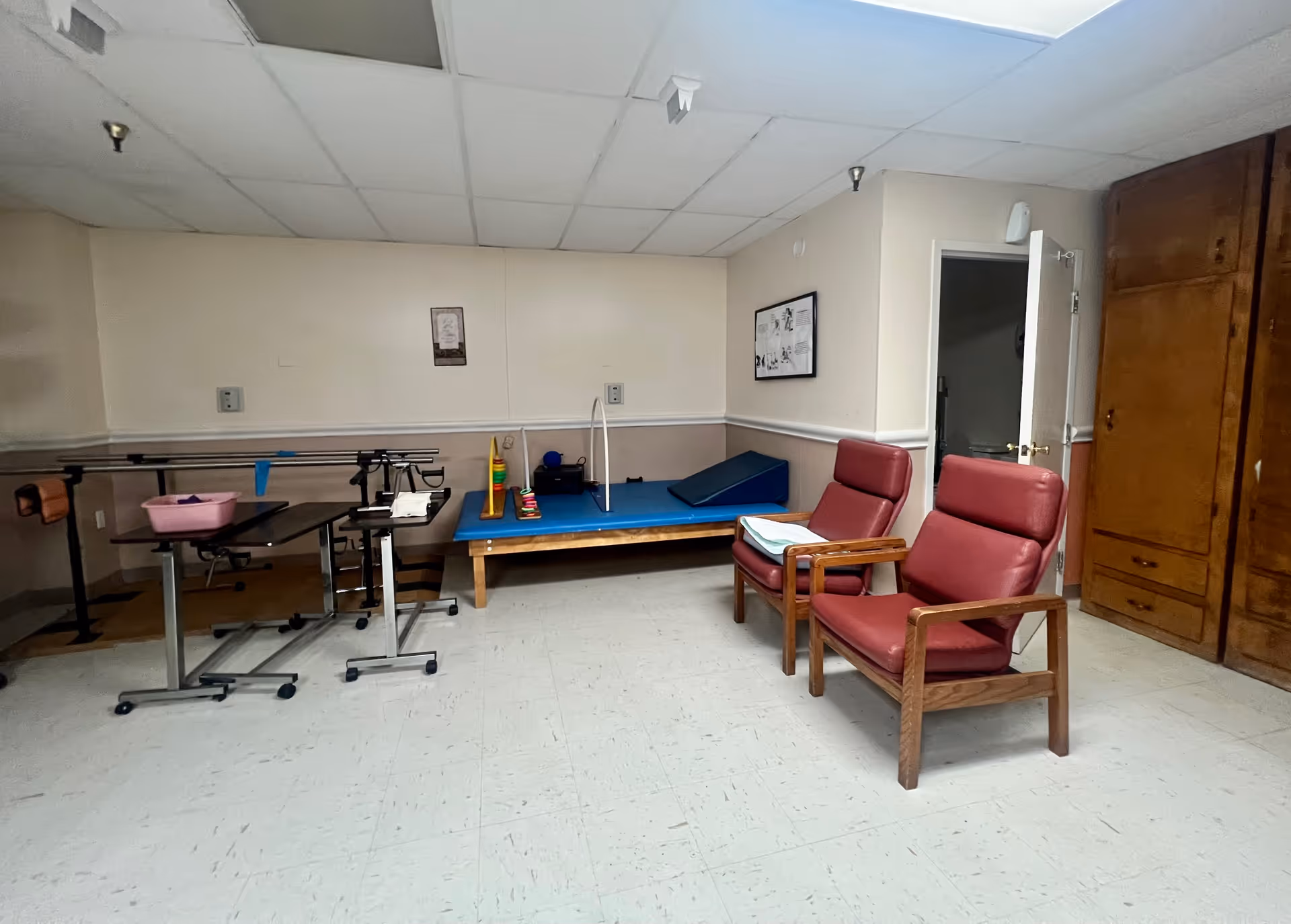 A therapy or rehabilitation room with two red cushioned chairs with wooden frames, a blue therapy mat with various therapy tools on it, parallel bars for walking assistance, a small table with a pink basin, and wooden cabinets against the wall. The room has a tiled floor and a drop ceiling with fluorescent lighting.