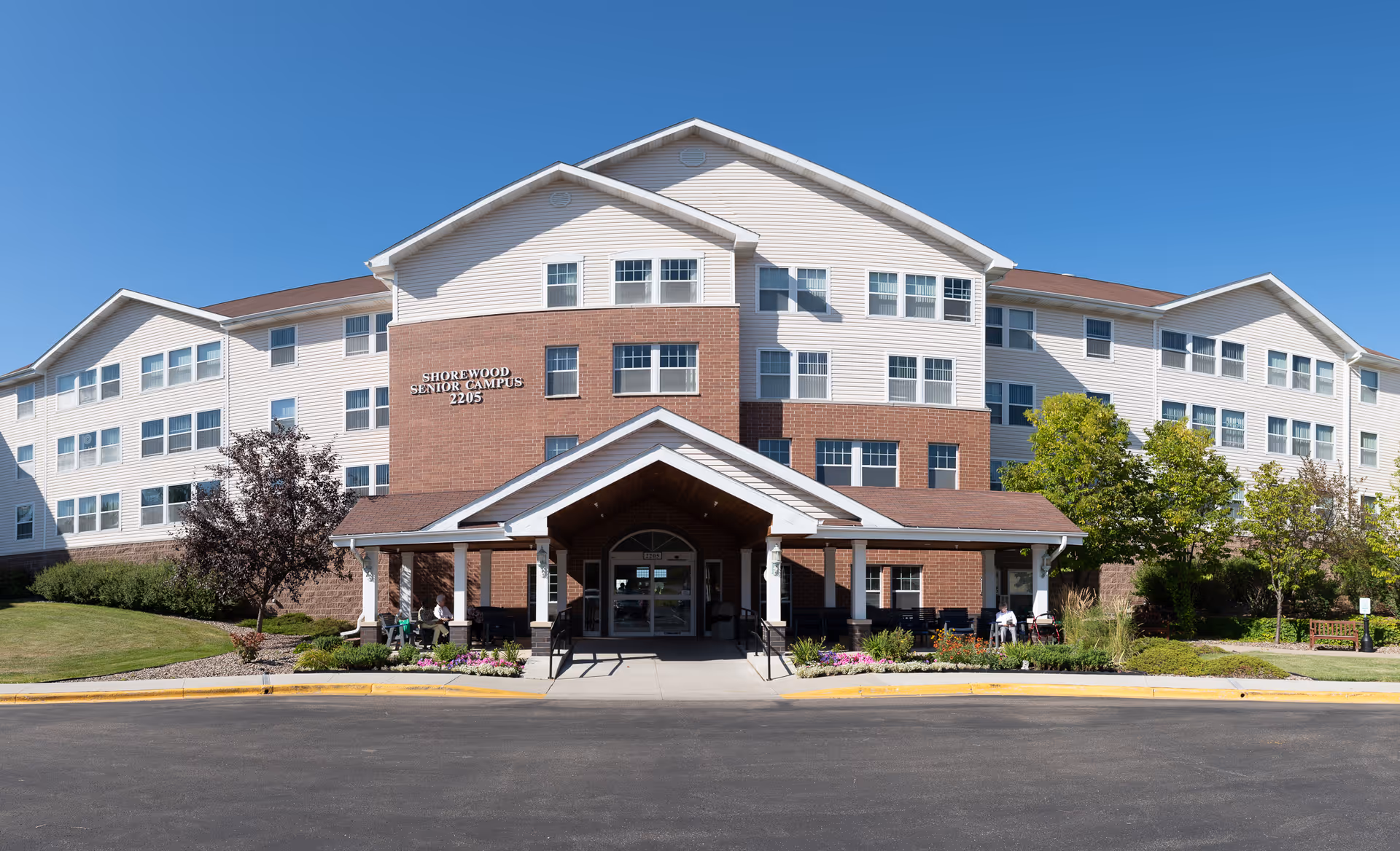 Front exterior view of Shorewood Senior Campus building with a covered entrance, multiple windows, and landscaped greenery under a clear blue sky.