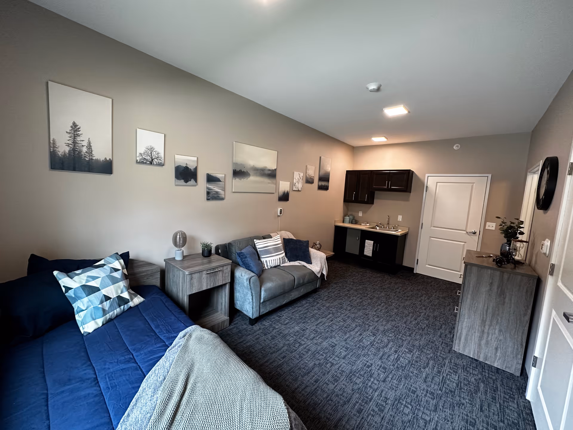 A cozy room with a single bed covered in blue bedding and a geometric patterned pillow. Next to the bed is a wooden nightstand with a small lamp and plant. A gray loveseat with multiple cushions and a throw blanket is positioned against the wall, which is decorated with a series of landscape and nature-themed framed pictures. At the far end of the room, there is a small kitchenette with dark cabinets, a sink, and a countertop. The floor is carpeted in a dark blue pattern, and the walls are painted a neutral beige color. A wooden dresser with decorative items and a wall clock are visible on the right side near the door.