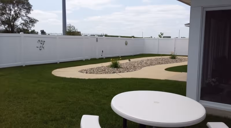 Outdoor area with a white round table and bench on green grass, a curved concrete pathway with decorative rocks and plants, and a white privacy fence in the background under a partly cloudy sky.