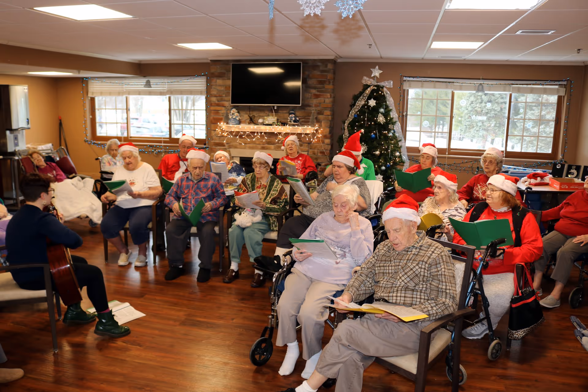 A group of elderly people wearing Santa hats sitting in a room decorated for Christmas, singing from songbooks while a person plays guitar. There is a Christmas tree with lights and ornaments, a fireplace with holiday decorations, and large windows showing a snowy outdoor scene.