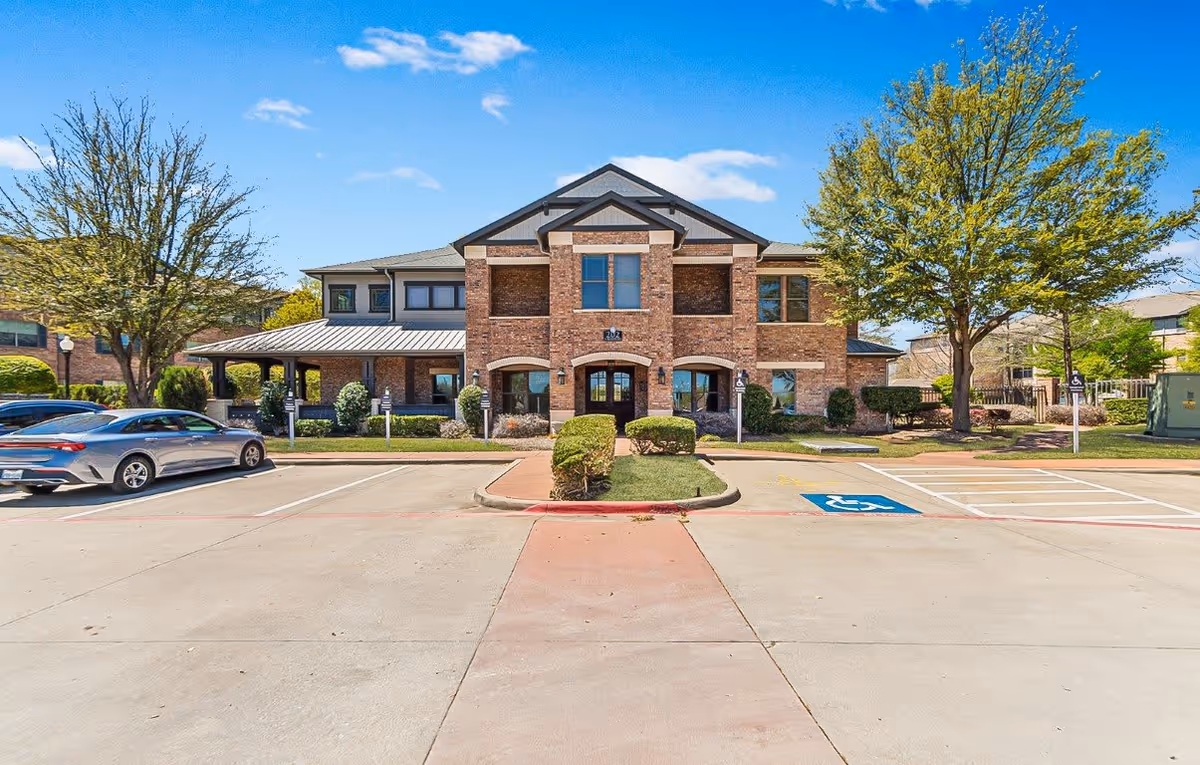 Front exterior view of a two-story brick building with a metal roof, surrounded by trees and landscaping. There is a parking lot in front with a few cars parked and a designated handicapped parking space visible. The sky is clear and blue.