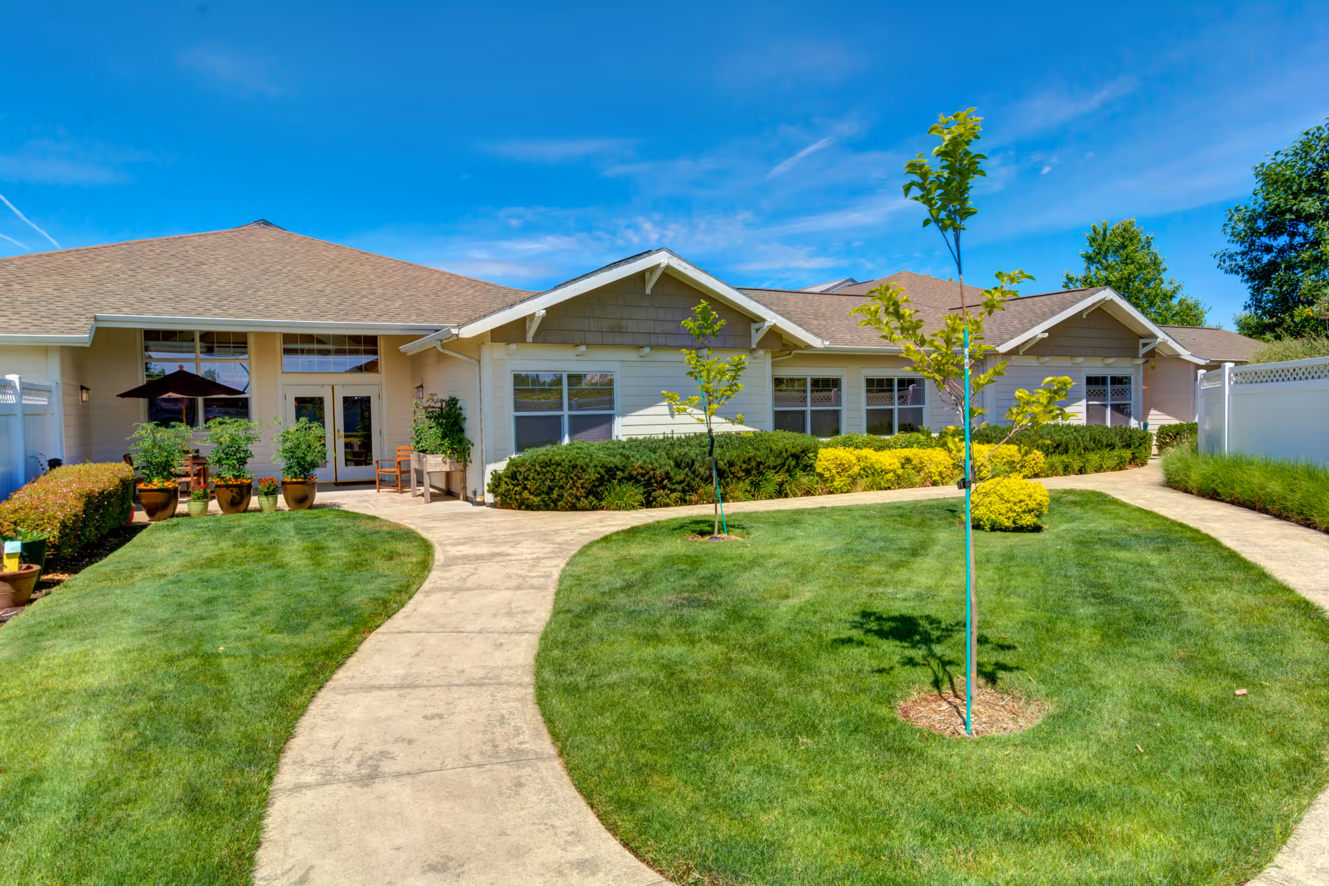 Exterior view of a single-story building with a beige roof and light-colored walls, surrounded by a well-maintained lawn with small trees and shrubs. A concrete pathway curves through the grass leading to a patio area with potted plants and outdoor furniture under a black umbrella. The sky is clear and blue.