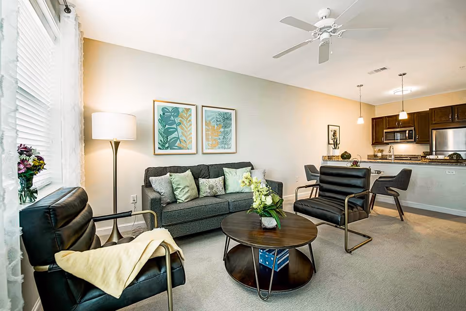 A modern living room with a gray sofa adorned with green and patterned cushions, two black leather armchairs with metal frames, a round wooden coffee table with a flower arrangement, a floor lamp, and two framed botanical prints on the wall. In the background, there is an open kitchen with dark wood cabinets, stainless steel appliances, and a breakfast bar with two gray chairs.