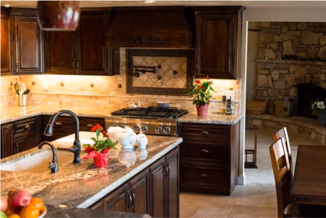 Open kitchen featuring a granite-topped island with sink, dark wood cabinetry, a stainless gas range, and a view into a stone fireplace area.