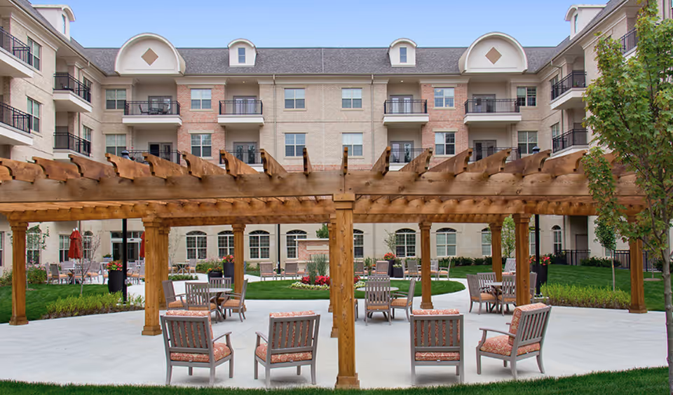 Courtyard patio with a wooden pergola, multiple outdoor chairs and tables, and a multi-story senior living building in the background.