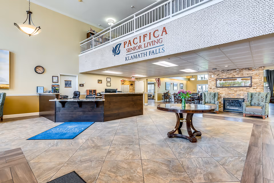 Spacious senior living lobby with a reception desk, central round table with flowers, seating area and a sign reading "Pacifica Senior Living Klamath Falls".