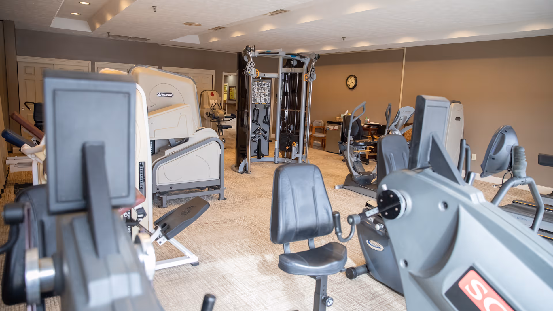 Interior view of a fitness room with various exercise equipment including stationary bikes, weight machines, and a multi-functional gym station. The room has beige walls, a carpeted floor, and a clock on the wall.
