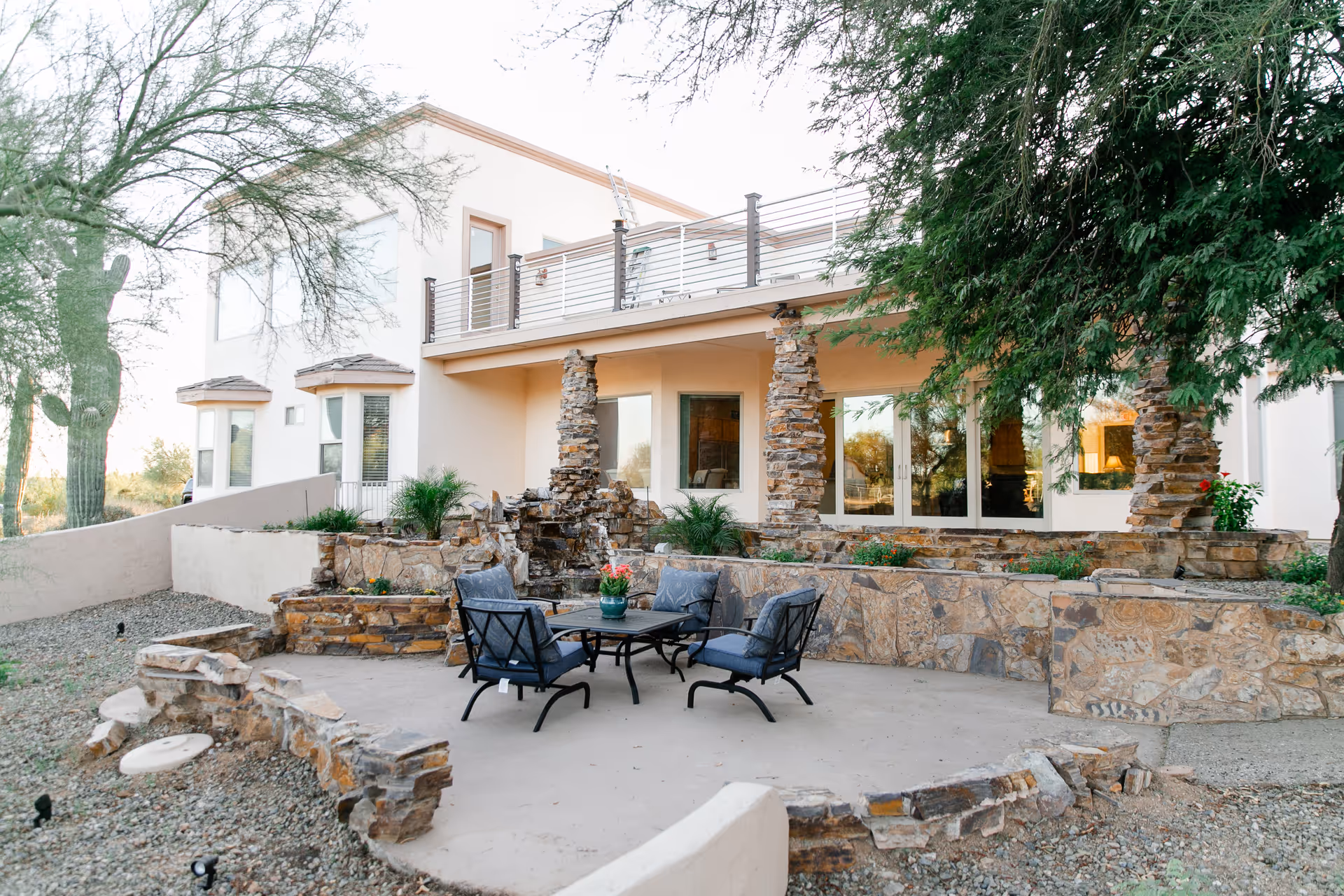 Outdoor patio area with stone walls and columns, featuring a seating arrangement with four cushioned chairs around a table. The patio is surrounded by trees and desert landscaping, and a two-story building with large windows and a balcony is visible in the background.