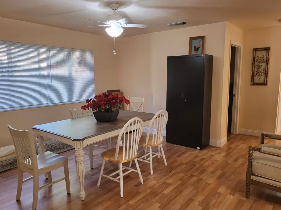 A dining area with a rectangular wooden table surrounded by five chairs, four white with wooden seats and one fully white. A black cabinet stands against the wall with a framed picture on top. The room has light-colored walls, a ceiling fan with a light, a large window with blinds, and wooden flooring. There is a floral arrangement in a black vase on the table and a framed floral picture on the wall near a doorway.