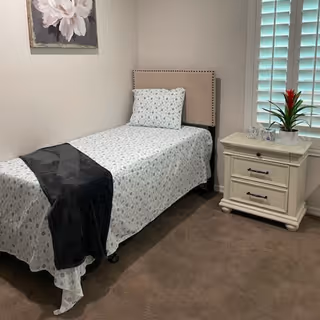 A small senior facility bedroom with a bed, nightstand, plant, and window blinds.