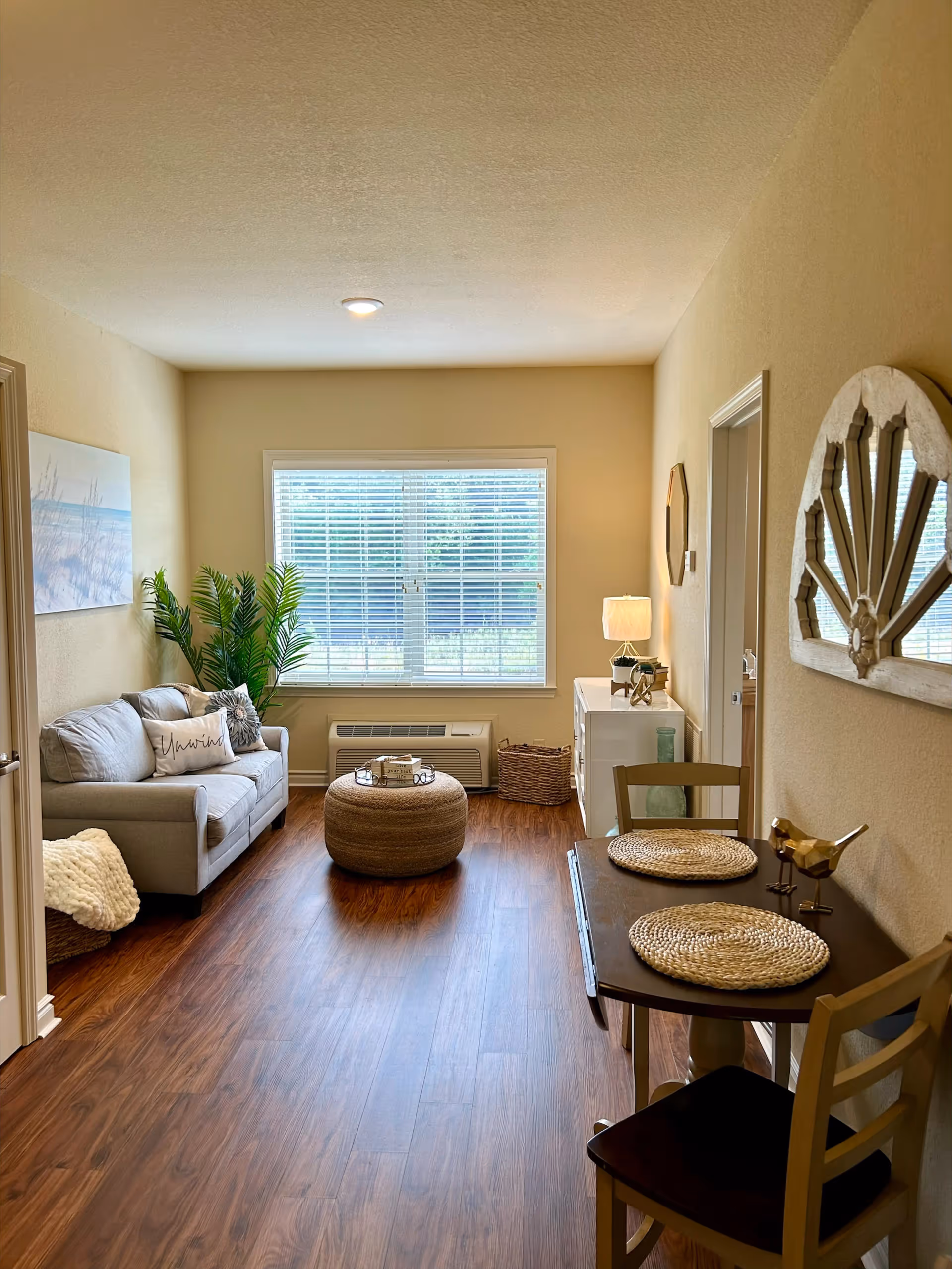 A cozy living area with a small gray sofa adorned with pillows, a round woven ottoman in the center, and a large window with blinds letting in natural light. To the right, there is a small dining table set with two chairs and placemats, decorated with two small bird figurines. A white cabinet with a lamp and decorative items sits against the far wall, and a large green plant is placed near the sofa. The room has wooden flooring and beige walls with a decorative mirror hanging on the right wall.