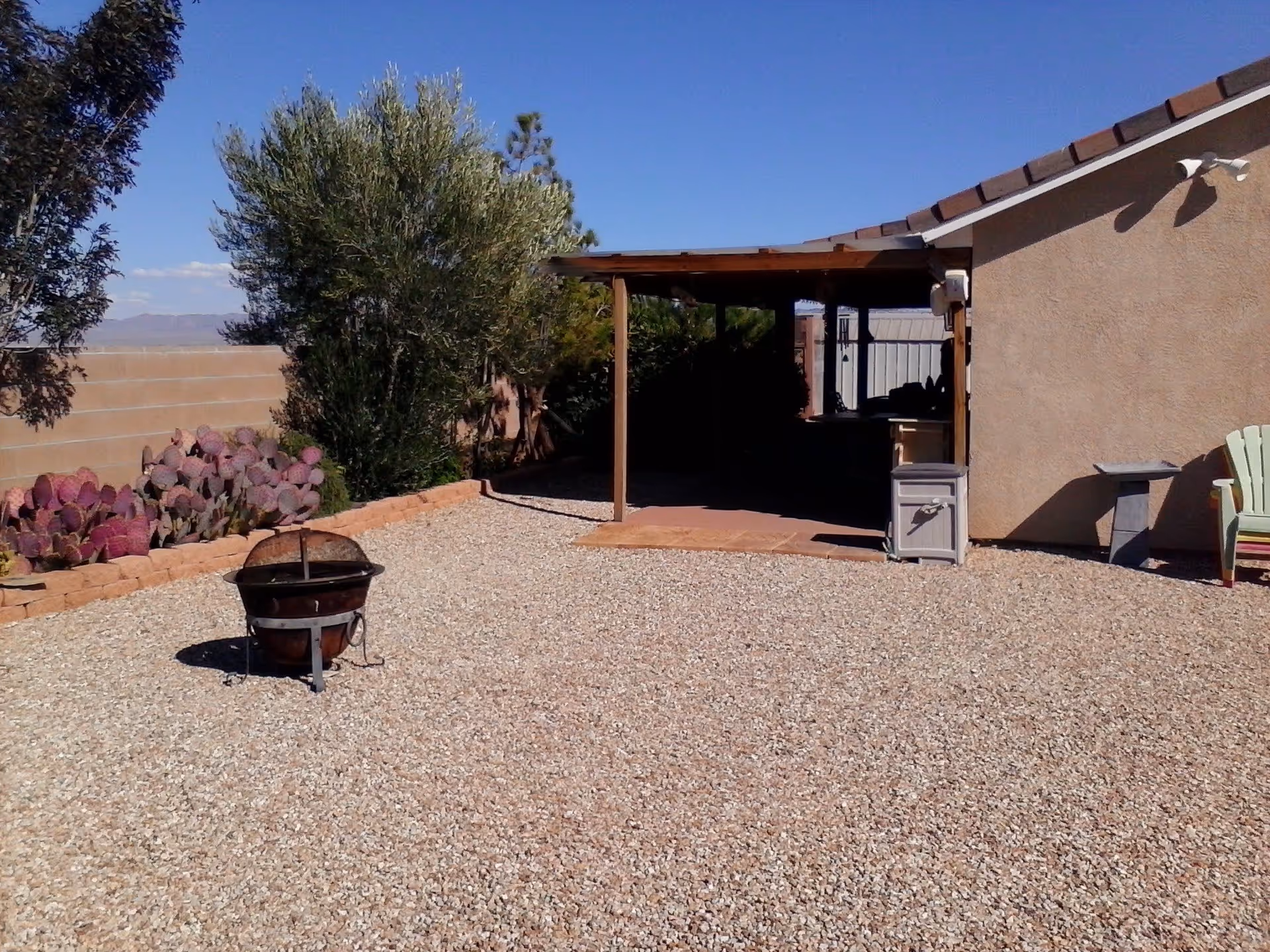 Outdoor area with gravel ground, a fire pit, various plants including cacti and trees, a covered patio attached to a building, and outdoor chairs.
