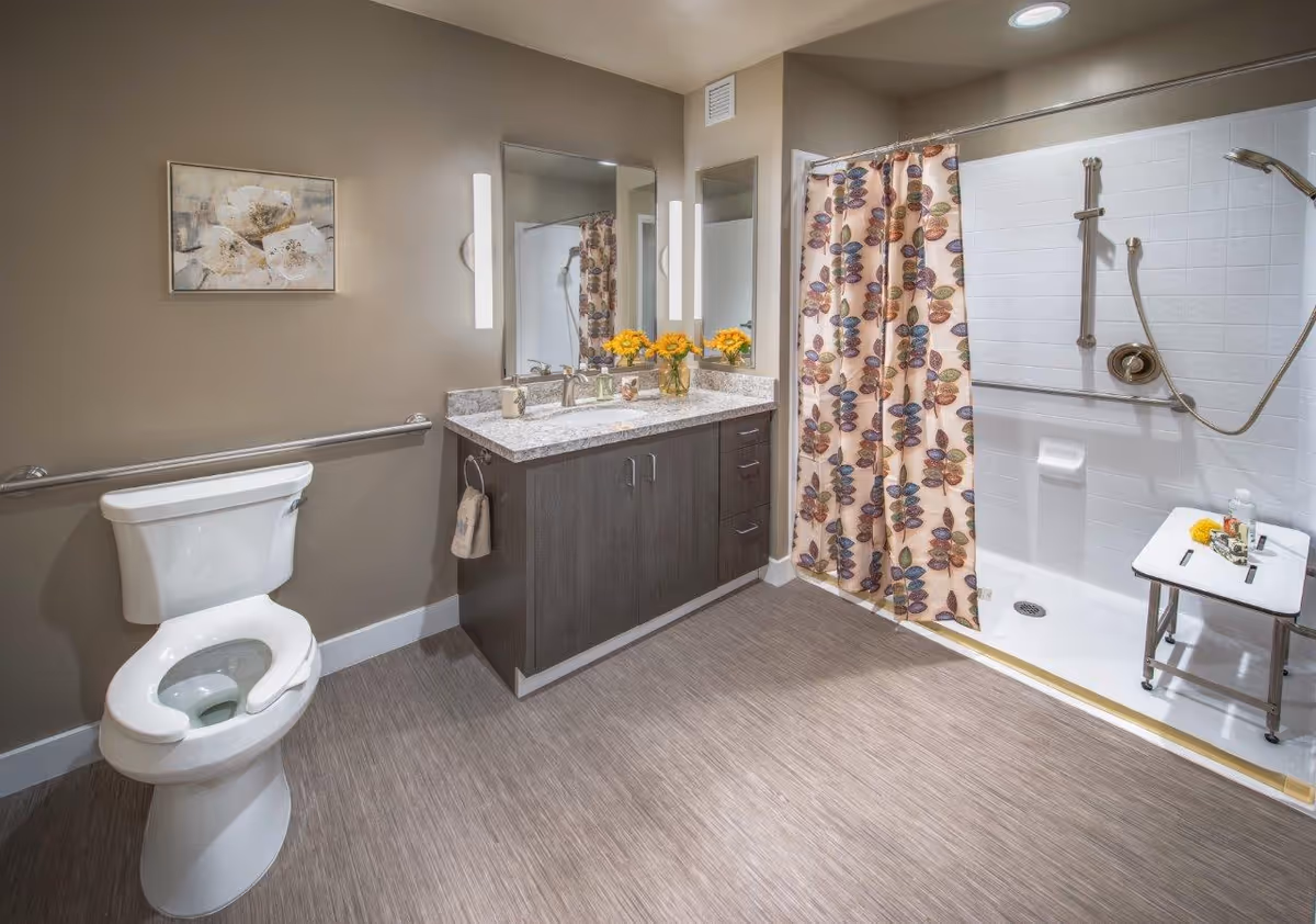 A clean and modern bathroom featuring a white toilet with a safety grab bar on the wall, a dark wood vanity with a granite countertop and a large mirror above it, and a walk-in shower with a floral-patterned shower curtain, a handheld showerhead, and a white shower chair. The walls are painted beige and there is a framed floral artwork hanging above the toilet.