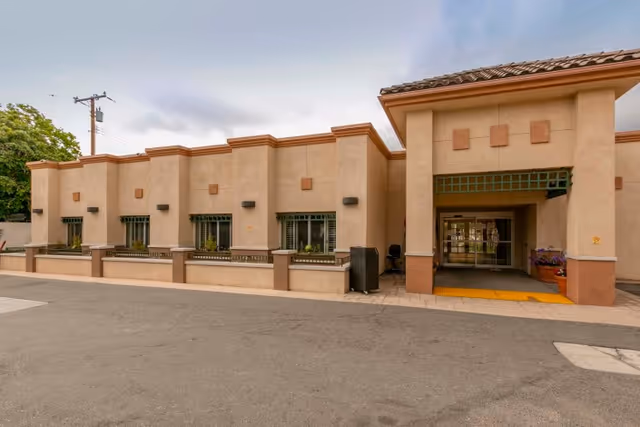 Exterior view of a beige stucco building with a tiled roof and several windows with green awnings. The entrance has a covered walkway with a green lattice design above it, and there are potted plants near the doorway. The area in front of the building is paved with asphalt.