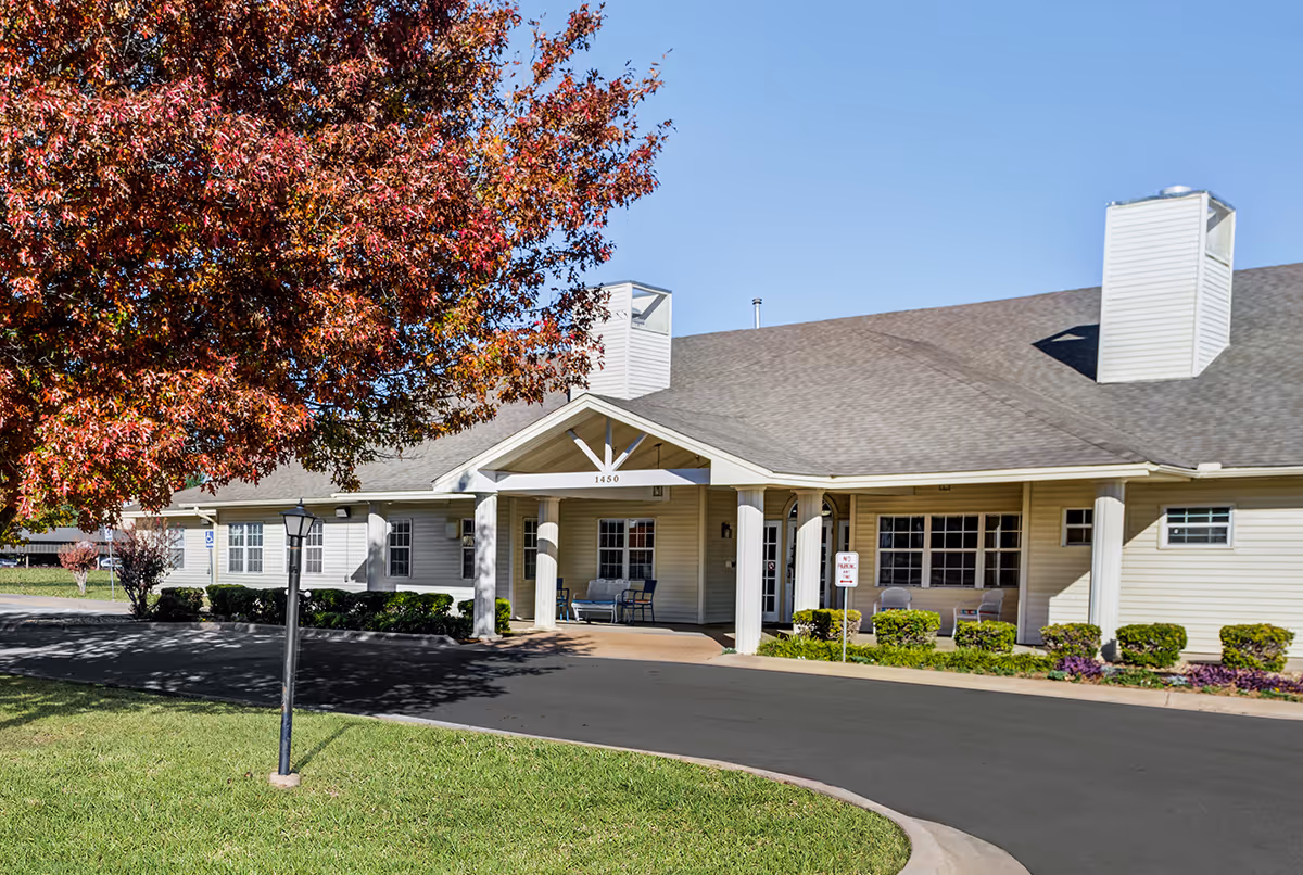 Exterior view of a single-story building with beige siding and a gray shingled roof under a clear blue sky. The entrance has a covered porch supported by white columns, with a few chairs placed outside. A tree with red and orange autumn leaves is visible on the left side, and a black lamp post stands on the grass near the driveway.